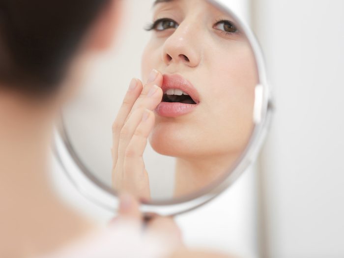 Woman with a cold sore looks in the mirror and touches her mouth