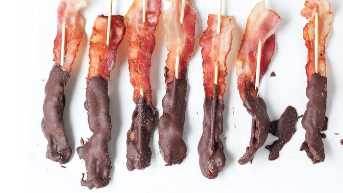 Skewers of bacon half dipped in chocolate, sitting on a white table, with chocolate spills