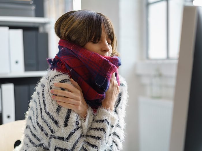 Cold weather, Woman shivers at her desk. She is bundled in a scarf and sweater and clutches her shoulders.