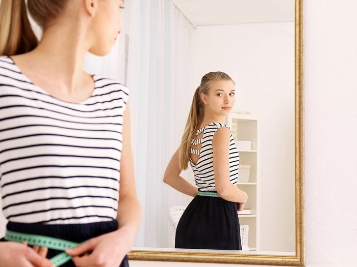Cold weather, Young woman looks in mirror and measures her waist, She wears a striped T-shirt and black skirt.