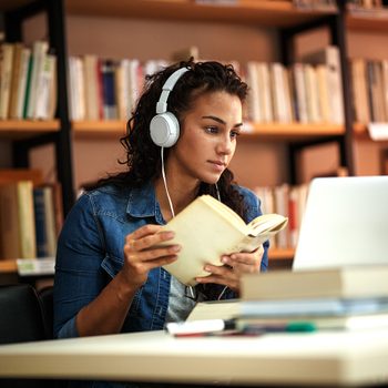 Cold weather, woman sitting at desk in library looking at laptop and listening on headphones. She also holds a notebook.