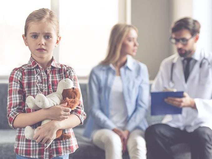 Dying, worried young girl holds her teddy bear as her mom talks to a doctor