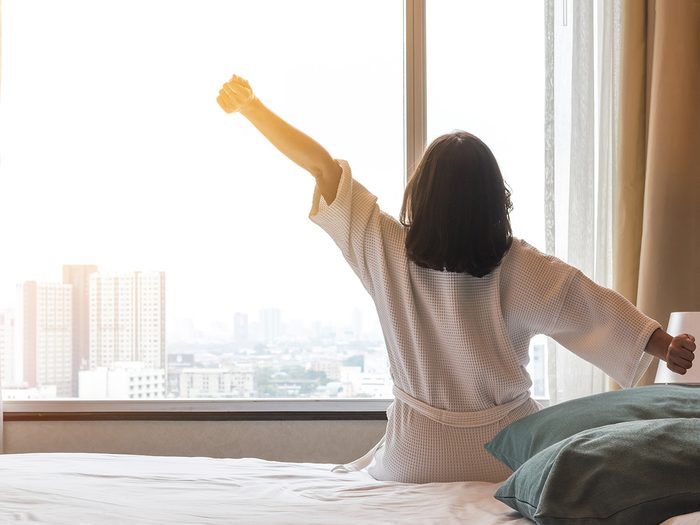 Skin care routine, Woman stretches as she gets out of bed and looks out the window at a city