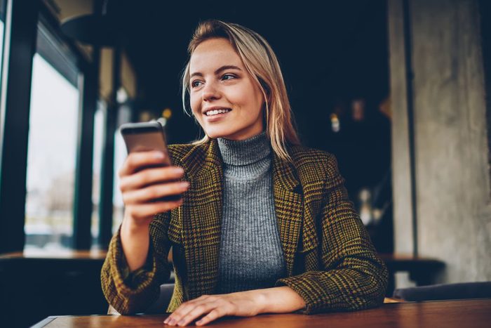Cheerful blonde female blogger in stylish jacket laughing and looking away while holding smartphone and messaging in chat via free 4G internet connection sitting at wooden table in coworking space