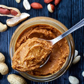 Jar and spoon of peanut butter and peanuts on dark wooden background from top view