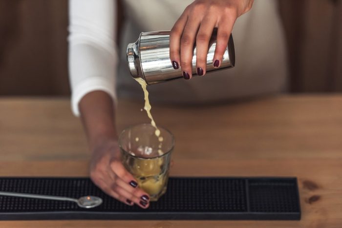 Cropped image of beautiful Afro American barista in apron preparing a cocktail in shaker while standing at bar counter