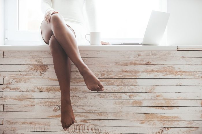 Beautiful seductress. Cropped image of gorgeous young African woman with beautiful legs sitting on the window sill with laptop laying near her 
