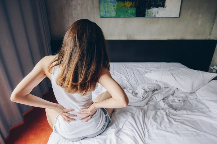 Back view of young woman suffering from Backache or Sore waist after her wake up in the bedroom. Conceptual of woman stretching suffering from sudden back pain.