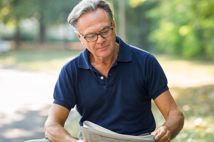 Senior man sitting at park and reading newspaper. Mature man wearing glasses while reading the finance section in a newspaper. Retired pensioner sitting in the park holding a newspaper.