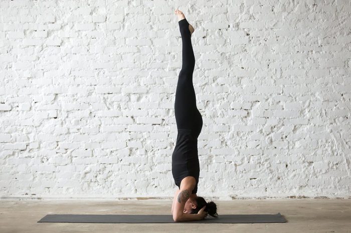 Young yogi attractive woman practicing yoga concept, standing in salamba sirsasana exercise, headstand pose, working out, wearing sportswear, black tank top and pants, full length, loft background