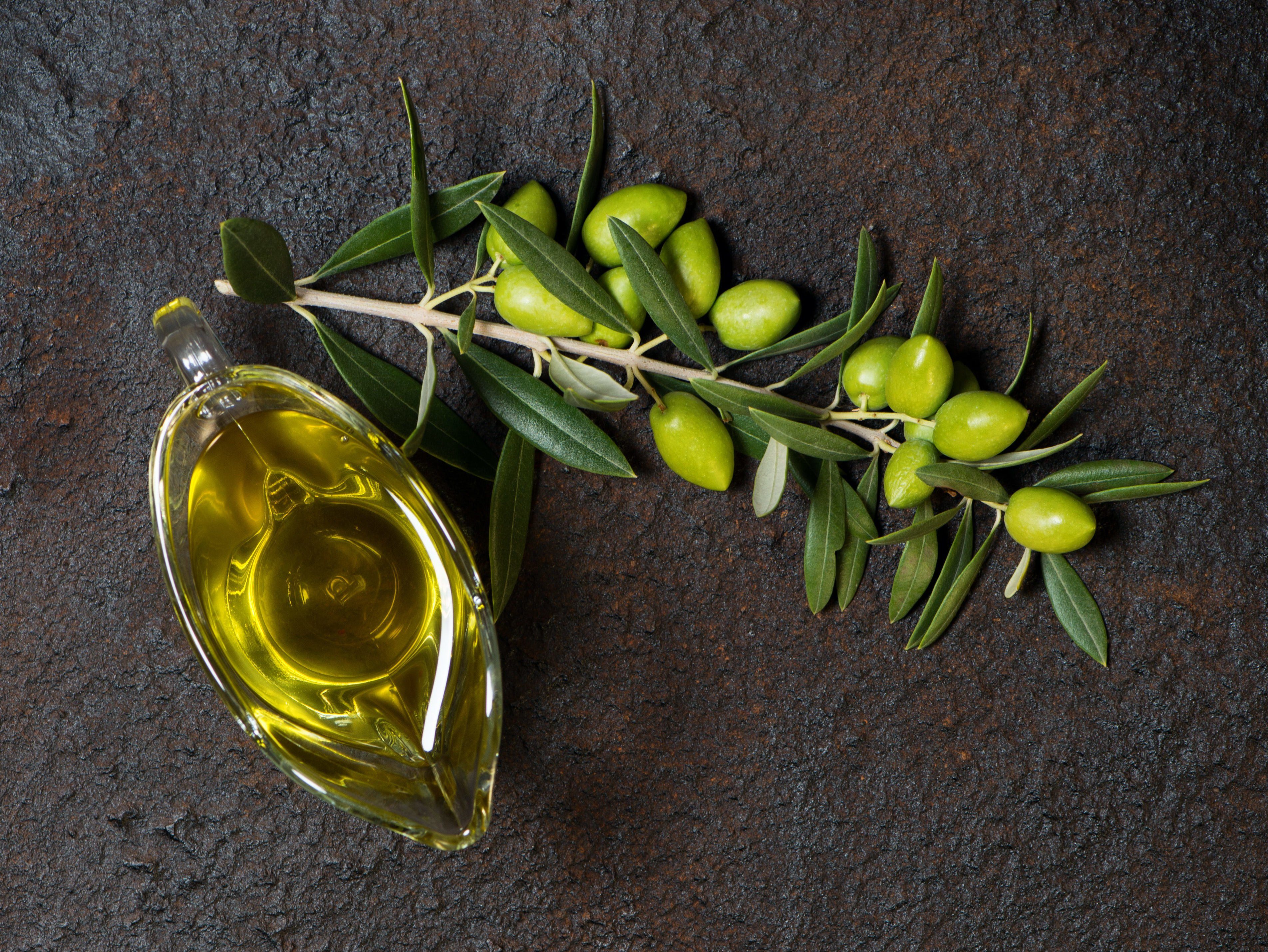 Top view of branch with green olives and glass sauceboat of olive oil on a black grunge metal background.