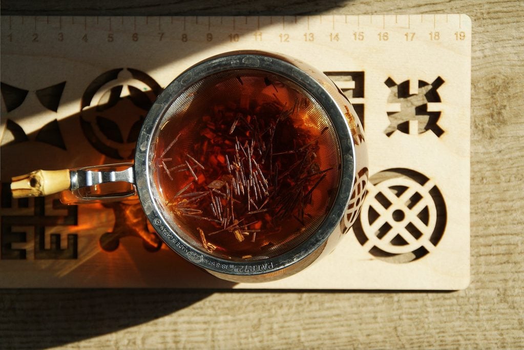 A glass cup with red-coloured wild Rooibos tea, wooden tea tray with geometrical shapes carved out, light brown pavement, sun and shadows.