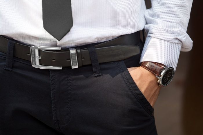 Closeup shot of male waist with hand in pocket dressed in black pants, belt, grey shirt, black tie and watch with brown watch strap. Formal wear.