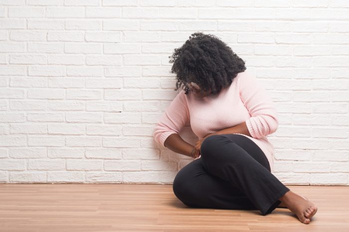 Young african american woman sitting on the floor at home with hand on stomach because nausea, painful disease feeling unwell. Ache concept.