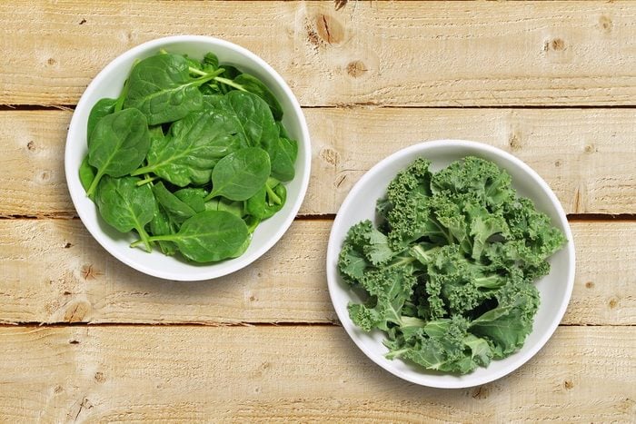 Two white bowls one containing spinach leaves and one containing chopped kale leaves. Shot from above on a rustic wooden background