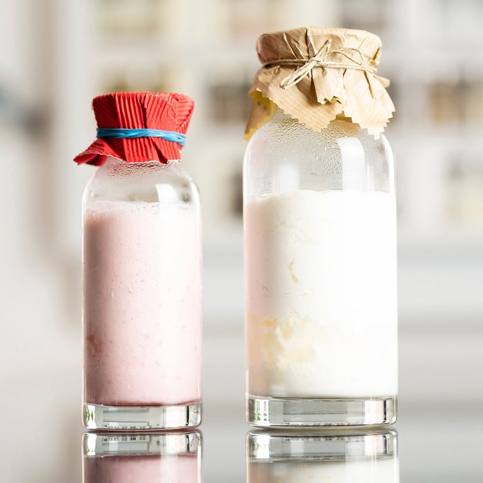 side view of glass bottles with a homemade raspberry smoothie with kefir yogurt and regular kefir yogurt with paper covers and kitchen shelf with spices in the background