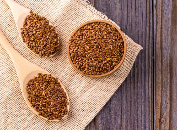 Brown flax seeds or flax seed in a small bowl on sacking and two wooden false on a brown wooden table photographed from above