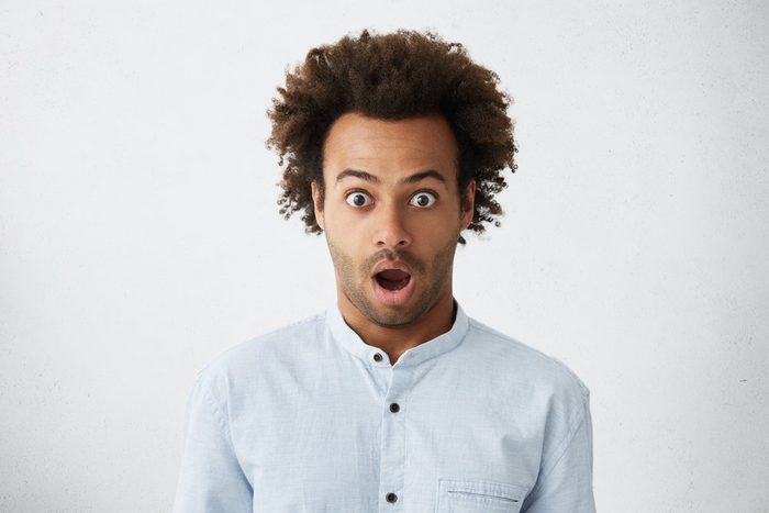 Studio portrait of bug-eyed dark-skinned man with bushy dark hair and bristle looking with surprise unexpecting to hear news. Scared man with wide opened eyes and mouth isolated over white wall