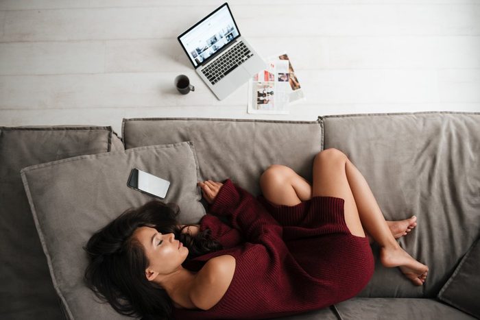 cause hemorrhoids | Top view of a beautiful asian woman in sweater sleeping on a couch at home with blank screen mobile phone and laptop