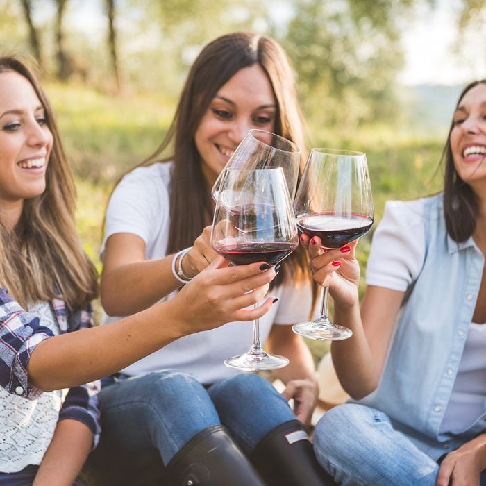Three happy women toasting with red wine while sitting with basket of grapes in the garden