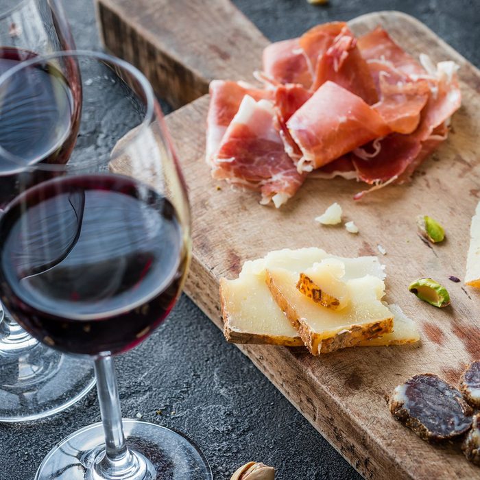 Platter with Spanish ham jamon serrano or Italian prosciutto crudo, sliced Italian hard cheese pecorino toscano, homemade dried meat salami, glasses of red wine and pistachios, on old wooden board