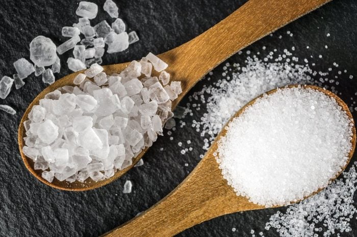 Different types of salt. Sea and kitchen salt. Top view on two wooden spoons on black background