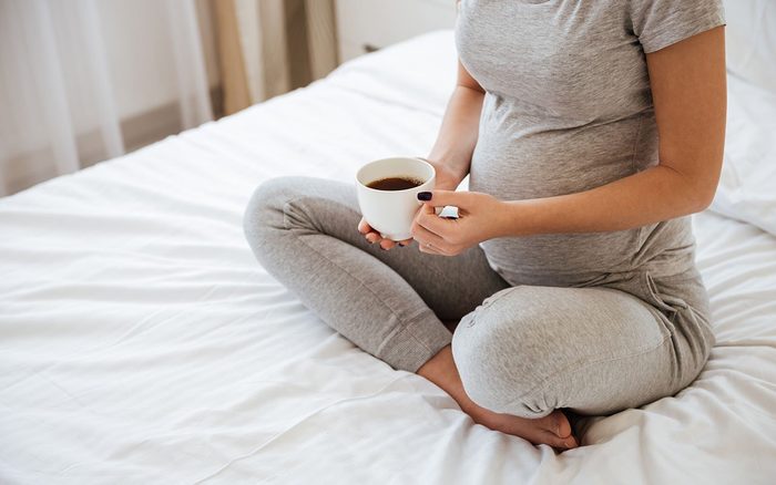 Closeup of pregnant young woman sitting on bed and drinking coffee at home