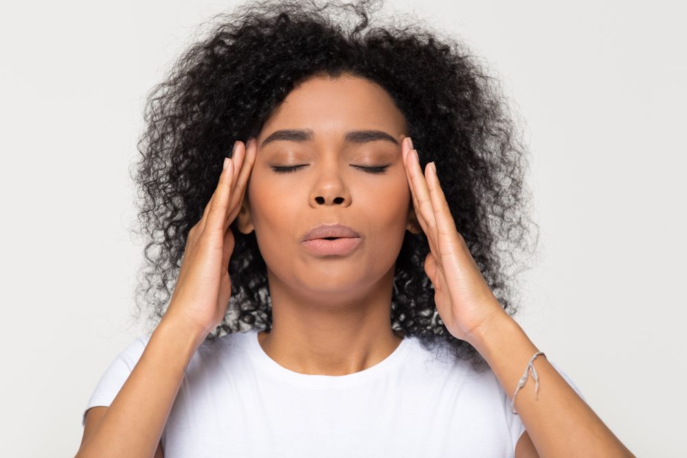 foods and drinks that cause migraines | Nervous african woman breathing calming down relieving headache or managing stress, black girl feeling stressed self-soothing massaging temples exhaling isolated on white grey studio blank background