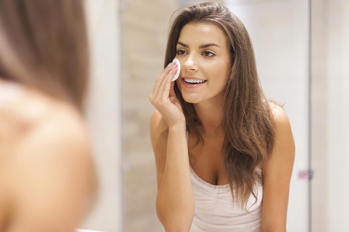 A woman applying a cleansing pad to her skin while standing in front of the mirror. | These skin care ingredients can cause breakouts.