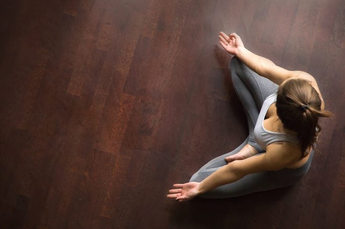 Young yogi woman practicing yoga, sitting in Half Lotus exercise, Ardha Padmasana pose, working out, wearing sportswear, grey pants, indoor, home interior, wooden floor, high angle view, copy space 