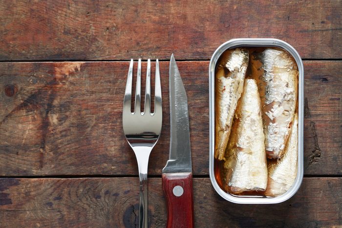 Open canned fish near fork and knife on wooden background with free space