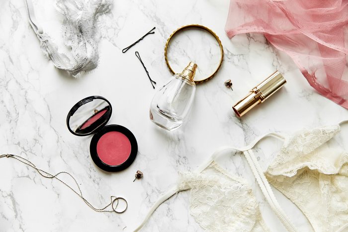 Overhead view of female lace lingerie, perfume, lipstick, blushes and jewelry items on white marble background. Top view, text space. Beauty routine concept