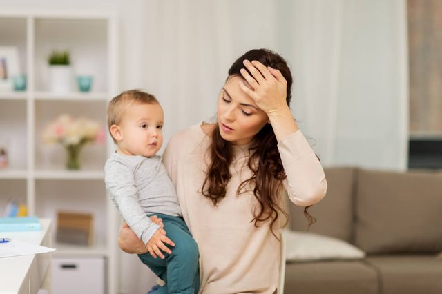 motherhood, multi-tasking and family concept - tired mother having headache with baby boy at home