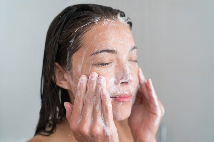 Skincare woman washing face in shower foaming facewash soap scrub on skin. Asian female adult cleaning body showering in hot water at home on in hotel as morning routine. Enjoying relaxing time.
