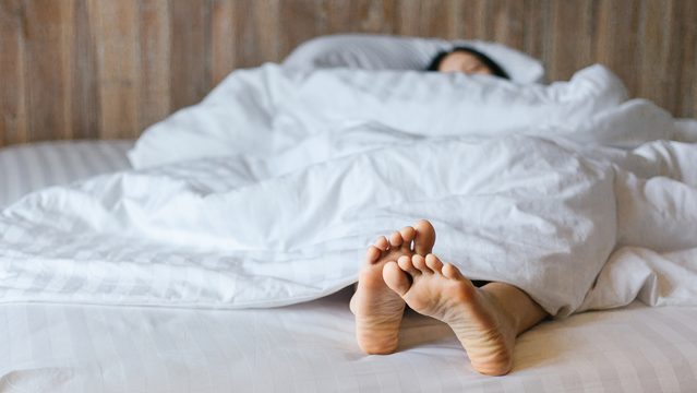 Fibromyalgia | Female feet under blanket flat lay. Female beautiful feet on the bed. Sleeping woman legs under white blanket