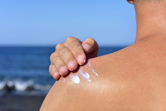 closeup of a young caucasian man seen from behind on the beach applying sunscreen to his own back