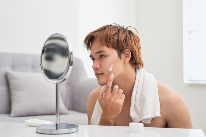 Handsome man looking at himself in mirror and applying cream lotion on face.