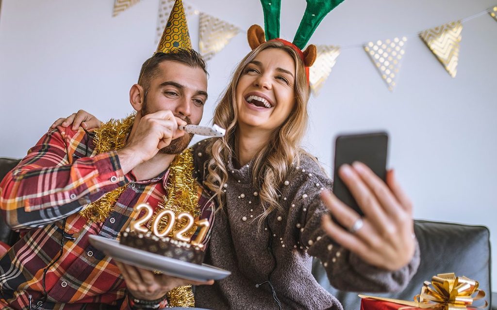 how to host a virtual new years party | Couple celebrating Christmas at home, holding chocolate cake with lit candles shaped as numbers 2021 and having a video call with friends