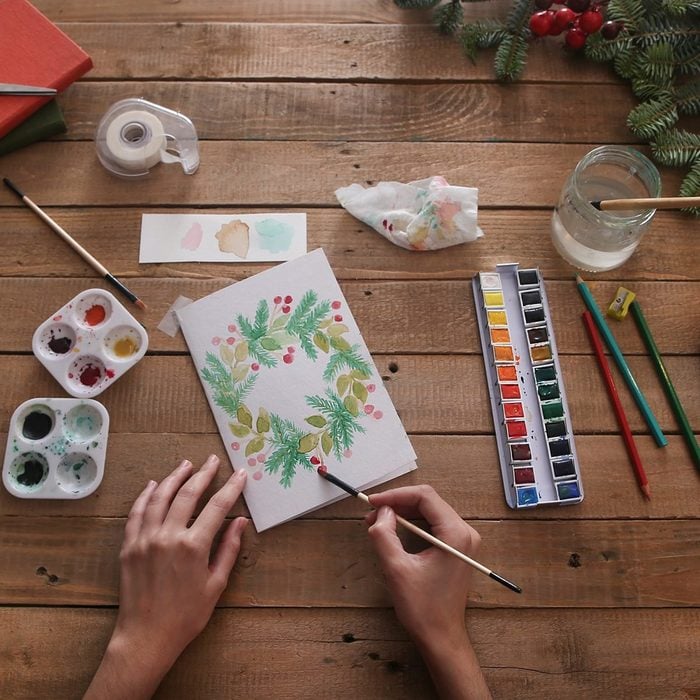 Young woman painting Christmas card with water colors, top view