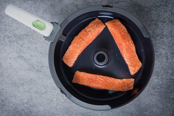 Three pieces of raw salmon in the air fryer at the center with a grey background. Air frying fish. Air frying salmon for a healthy salmon meal. Uncooked salmon in a pan shot in the center.