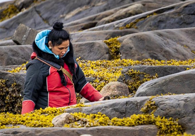 Bernice Clarke wears a red jacket and walks outside with a background of rocks and lichen